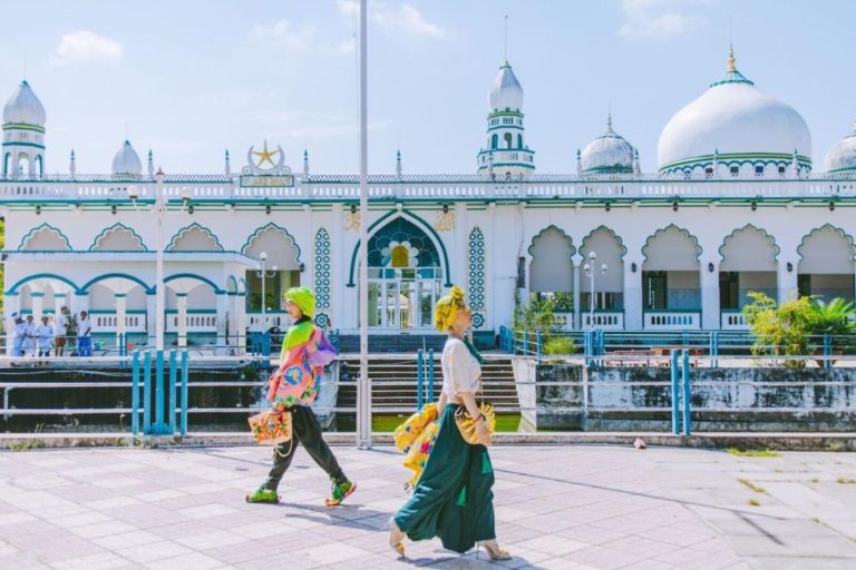 Two women in colourful headscarves and dresses walking past a white mosque with domes.