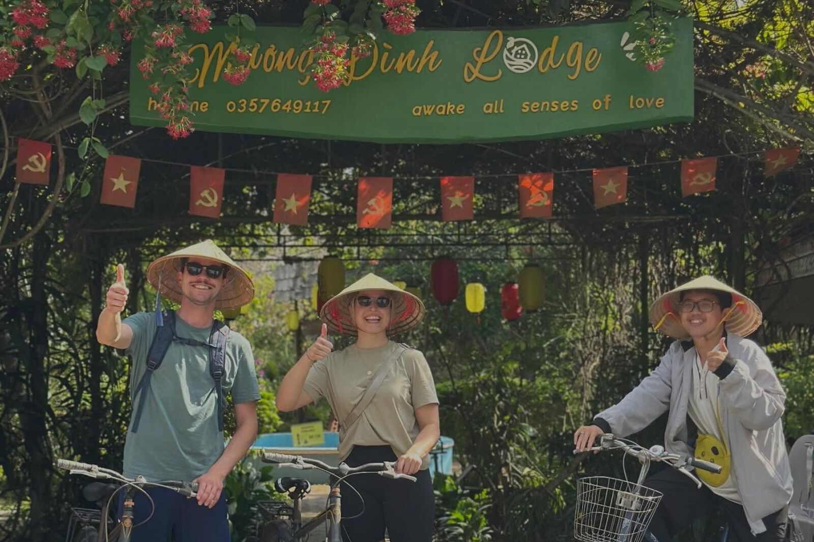 Three guests in conical hats with bicycles under the Mương Đình Lodge gate sign and red flags.