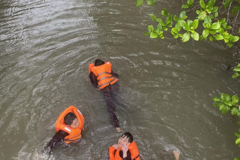 Children in orange life jackets swimming in a brown canal.
