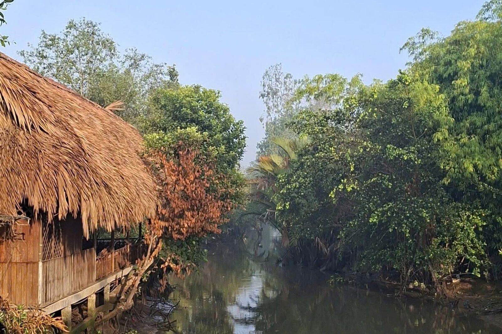 A thatched hut beside a quiet canal lined with trees.