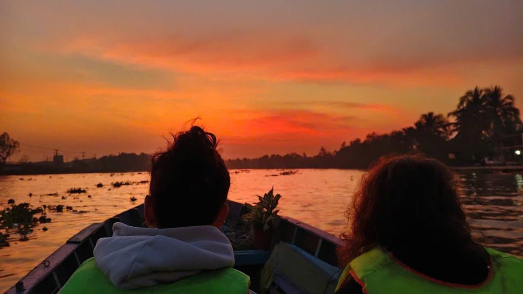 Guests in a boat facing a fiery orange sky over the river.