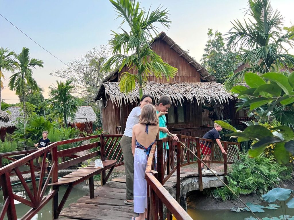 Guests on a wooden bridge by a thatched cottage, one holding a fishing pole.