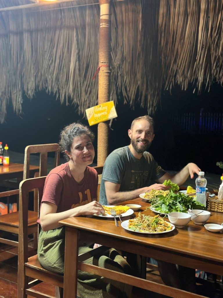 Two guests smiling at a table set with dishes under a thatched roof.