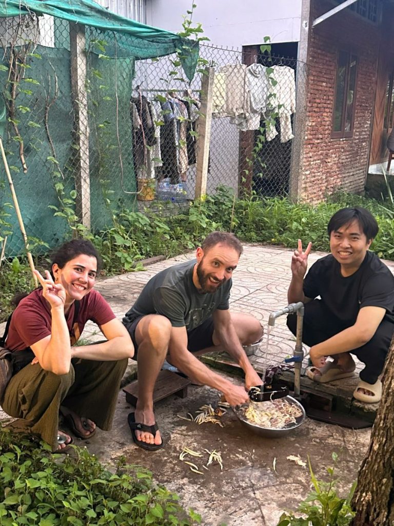 Three people squatting by an outdoor tap in a yard, one slicing pale strips into a steel basin.