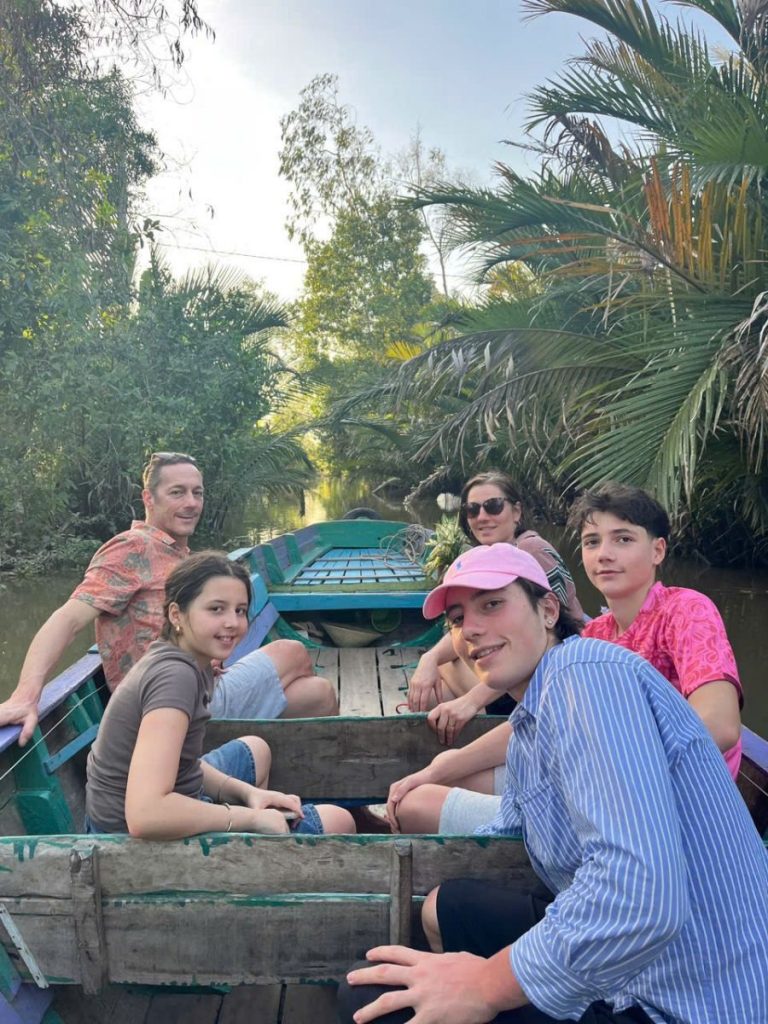 A family smiling in a wooden boat on a canal lined with water palms.