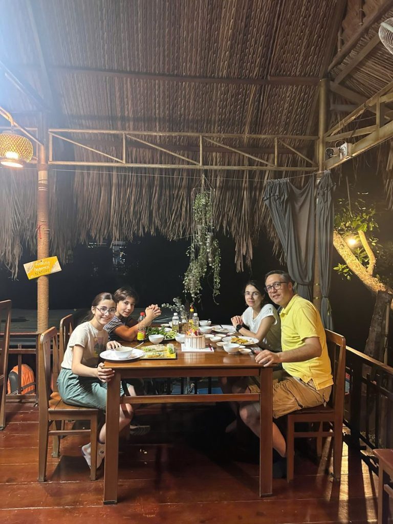 A family dining at a wooden table under a thatched roof in the evening.