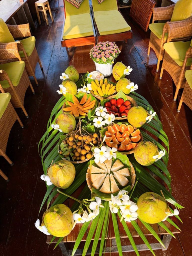 A long table on a boat laid with fruit, coconuts and white flowers on palm leaves.