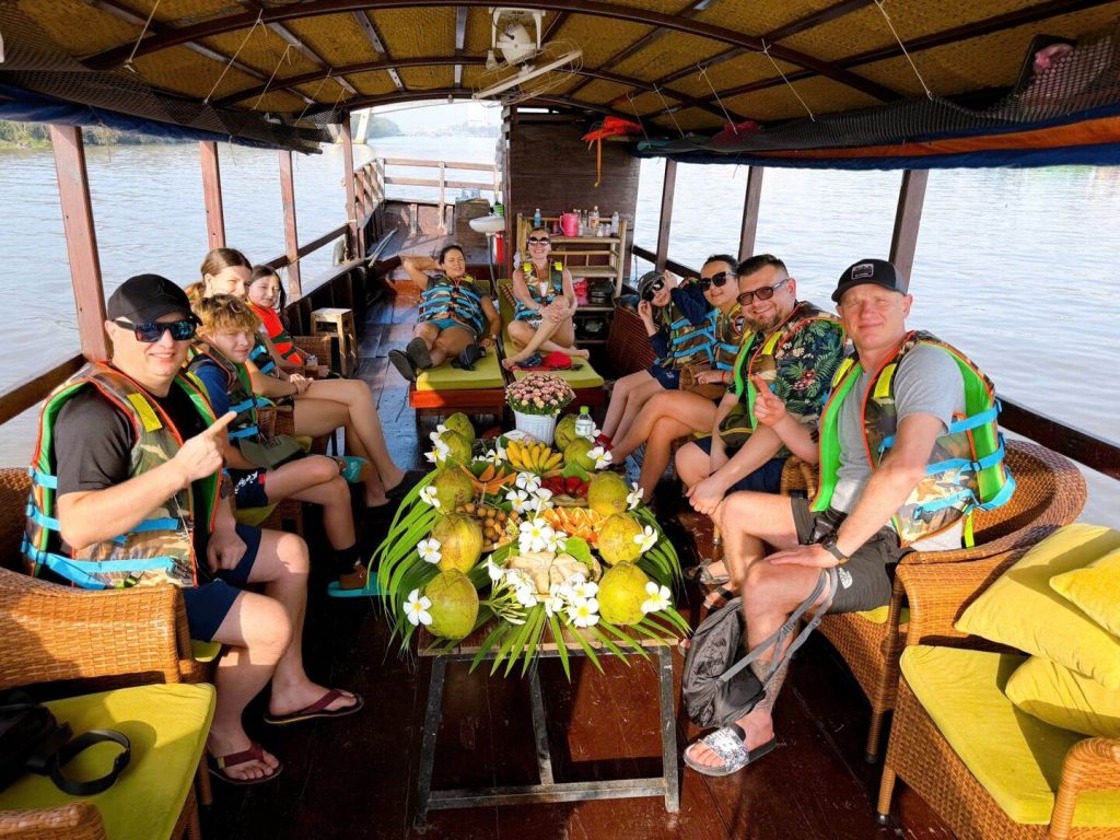 Du khách mặc áo phao ngồi quanh bàn trái cây trên chiếc thuyền có mái che.