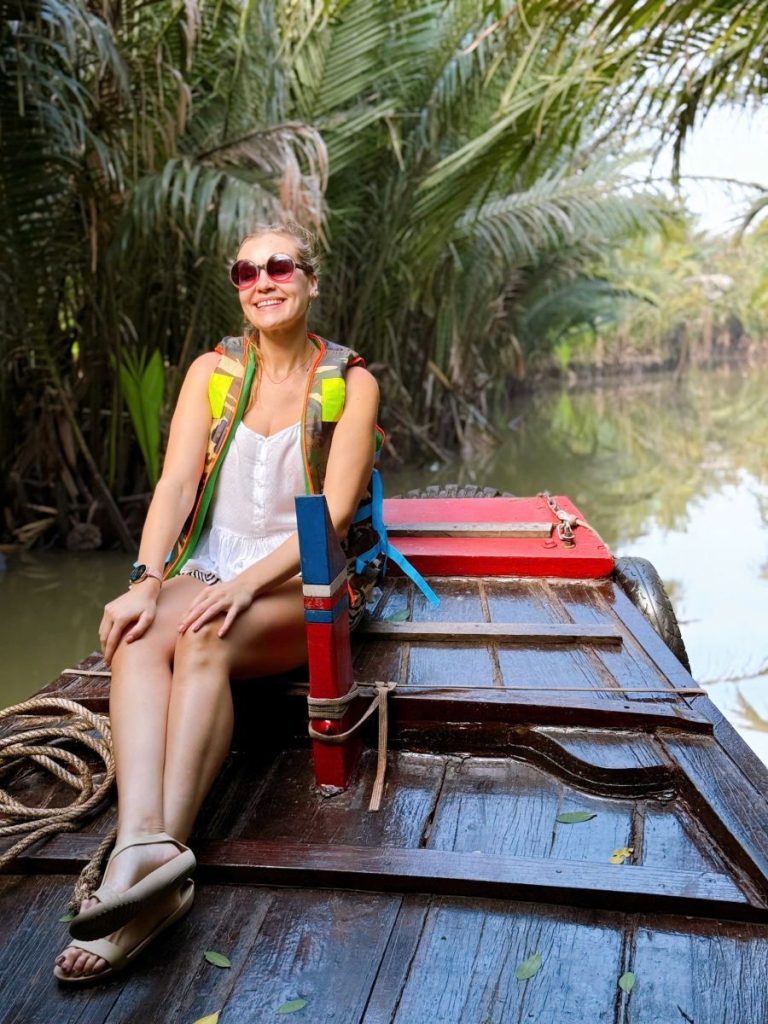 A smiling woman in sunglasses at the bow of a wooden boat among water palms.