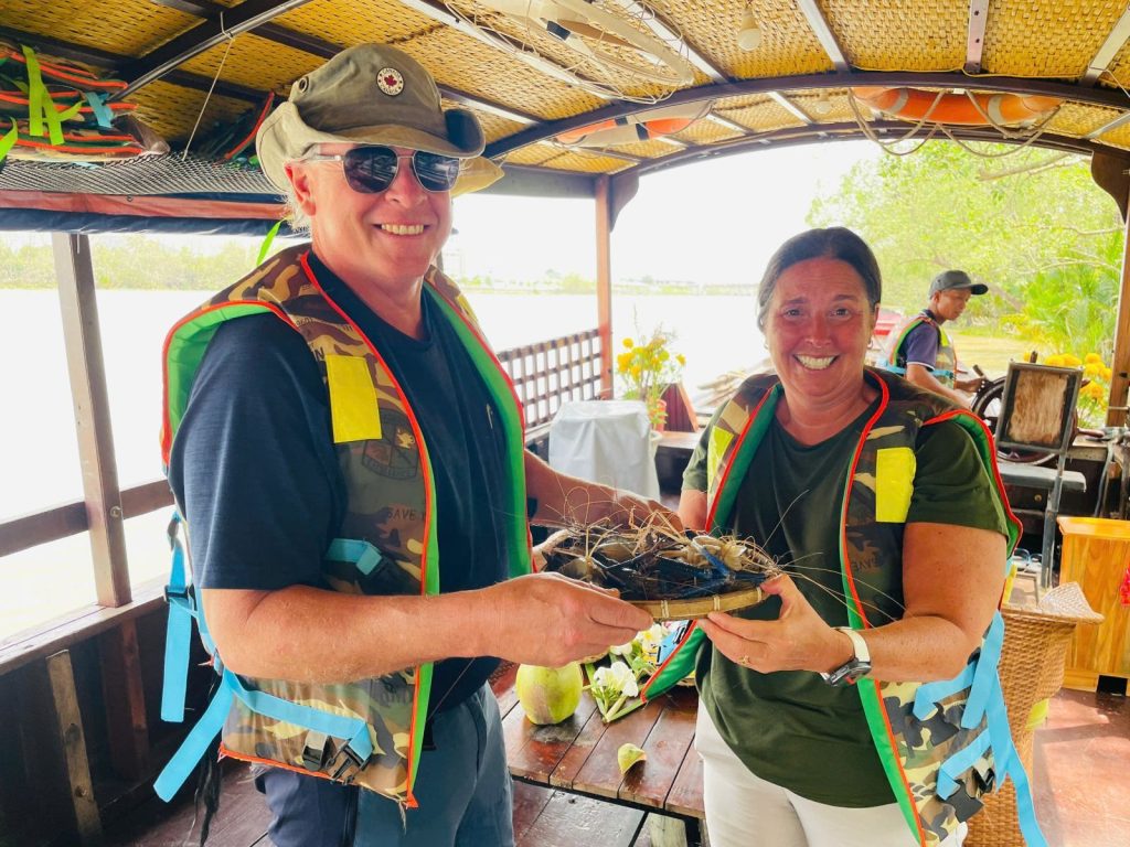 Two guests in life jackets on a covered boat, holding a small tray of fresh catch tied with string.