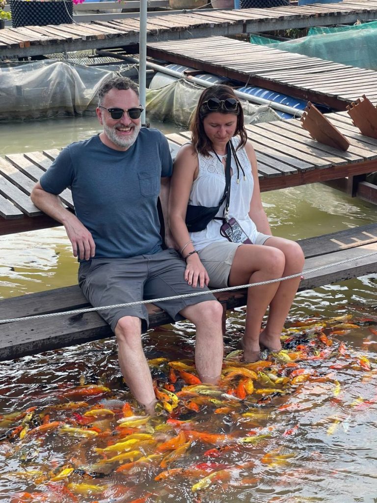 Two guests on a wooden platform dangling their feet among a crowd of orange fish.