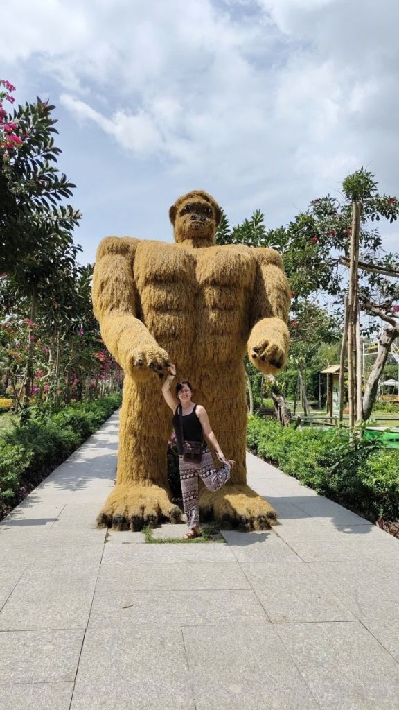 A woman posing beside a large straw gorilla figure on a paved path.