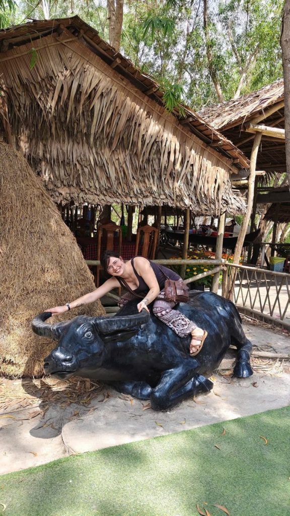 A woman sitting on a black water buffalo statue beside a thatched hut.