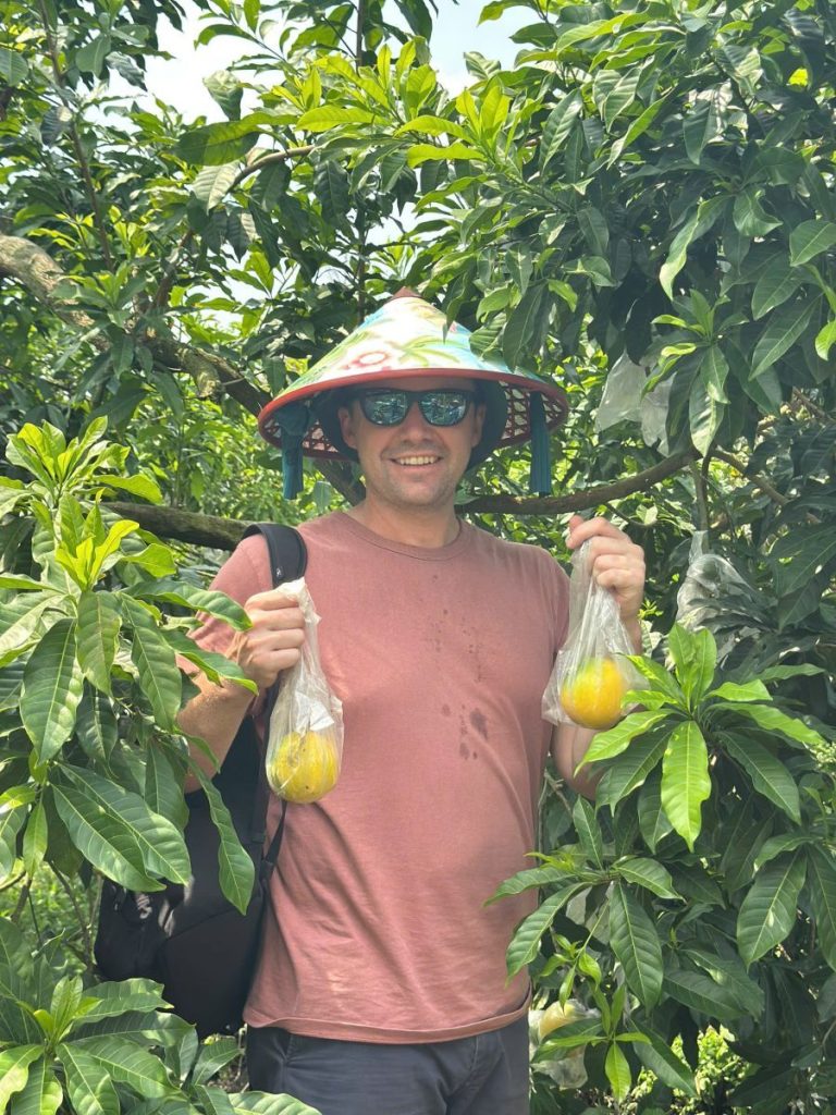 A smiling man in a conical hat holding up two bags of fruit in an orchard.