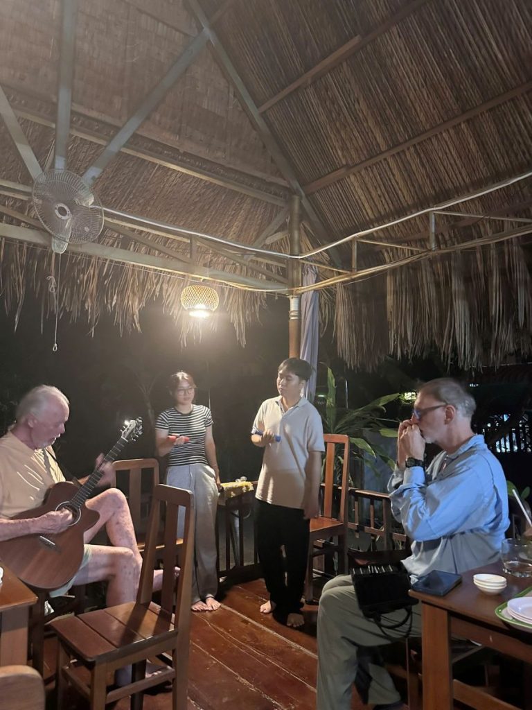 Guests chatting and playing guitar under a thatched pavilion in the evening.