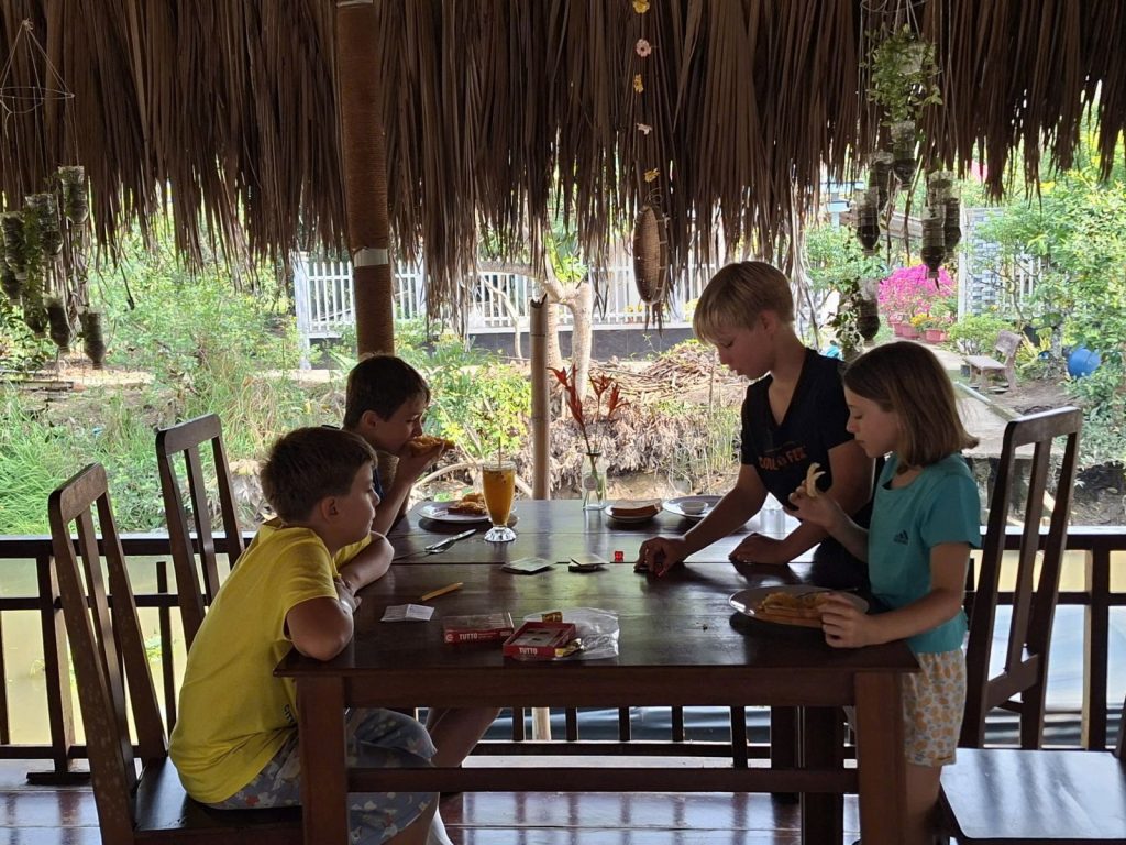Four children playing a game at a table in a thatched pavilion.
