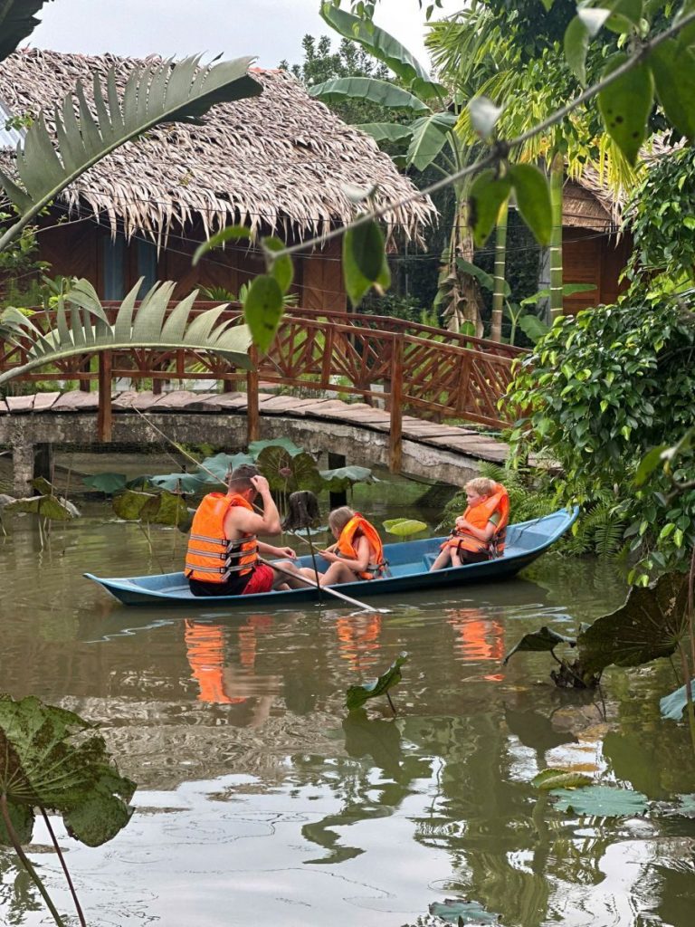 Guests in life jackets paddling a small boat under a wooden bridge by a thatched cottage.