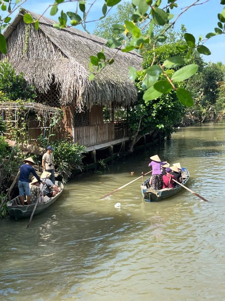 Rowers in conical hats paddling guests along the canal past a thatched stilt house.