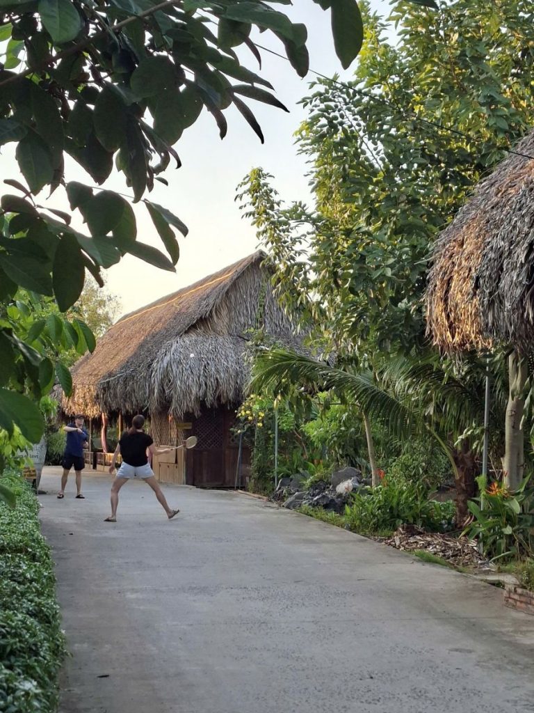 Two people playing badminton on a concrete lane lined with thatched huts and trees.