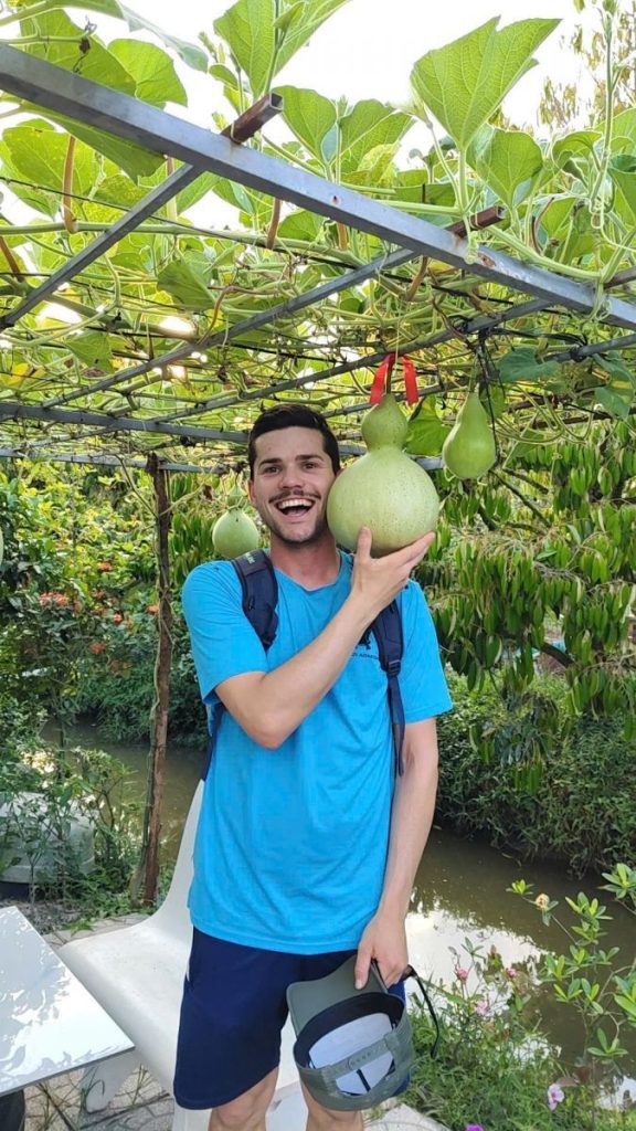 A smiling man holding a large green fruit under a trellis hung with more.