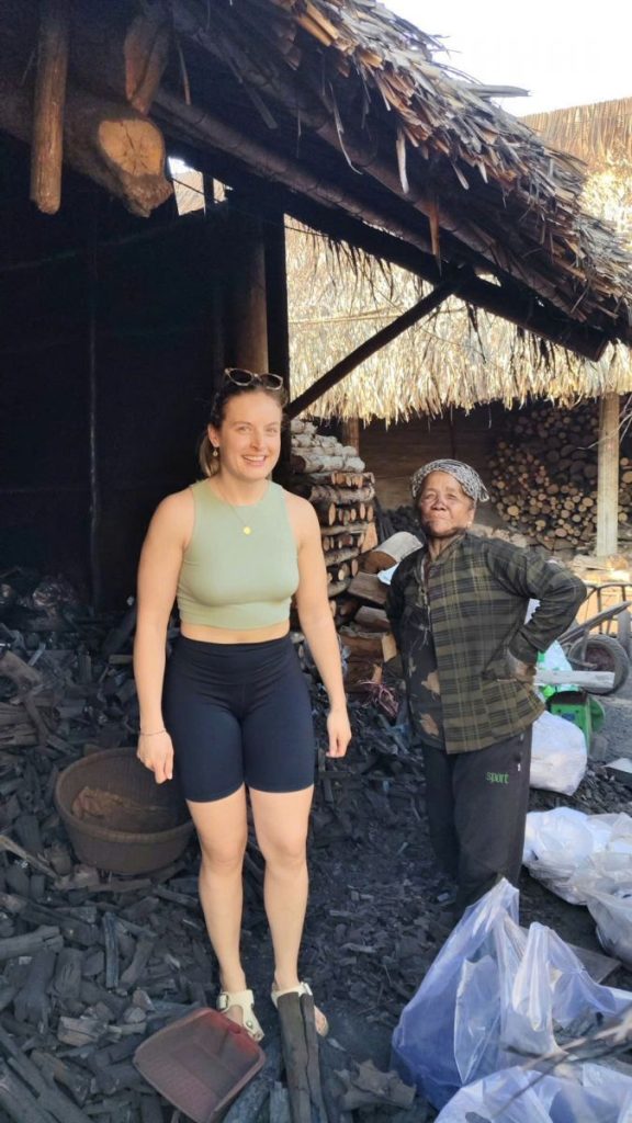 A visitor and an elderly local woman by a thatched shed piled with black charcoal.