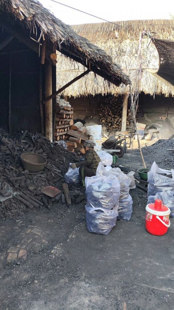 Bags of charcoal and a red bucket outside a thatched shed.