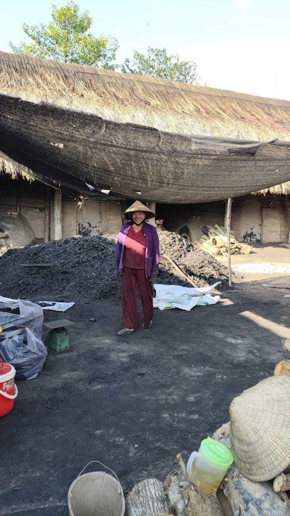 A woman in a conical hat standing in a yard of charcoal under a thatched roof.