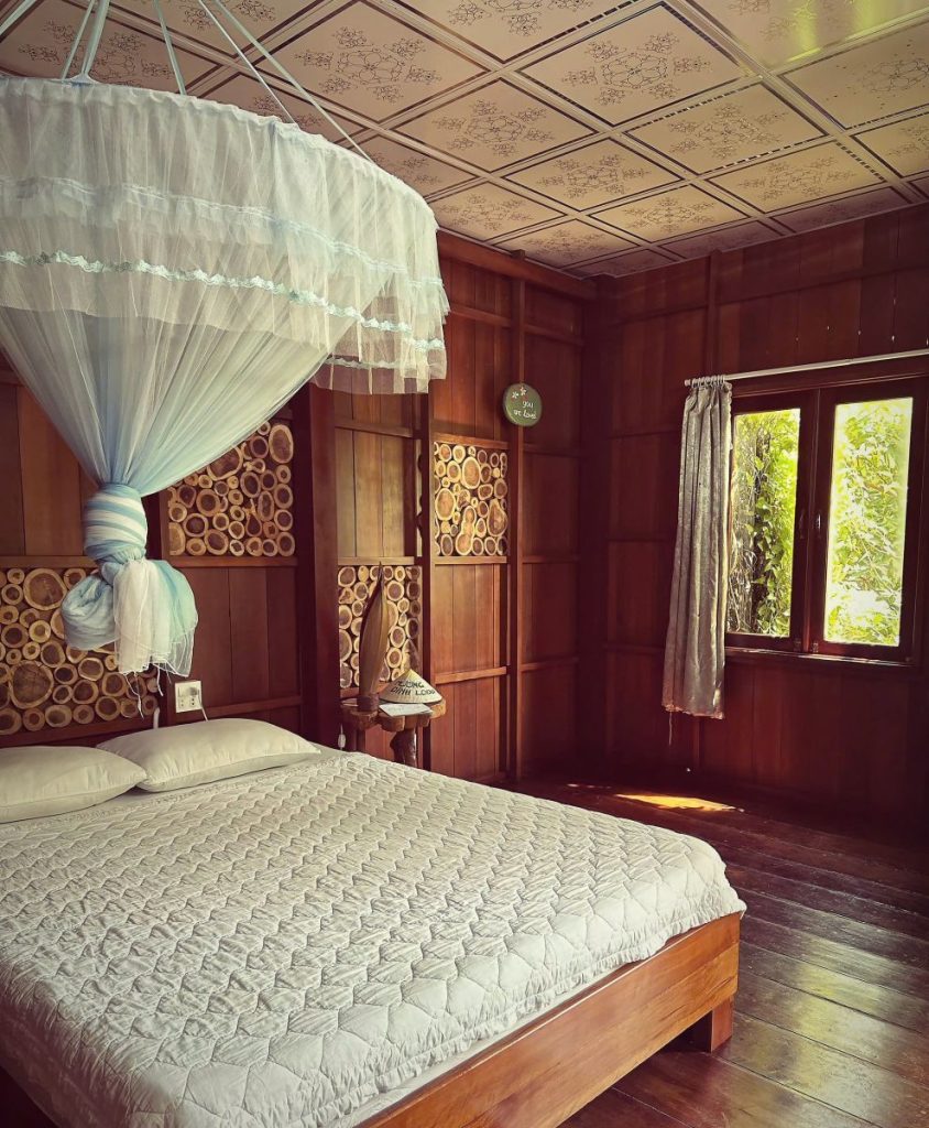 A wooden room with a bed under a round net canopy and a window onto greenery.
