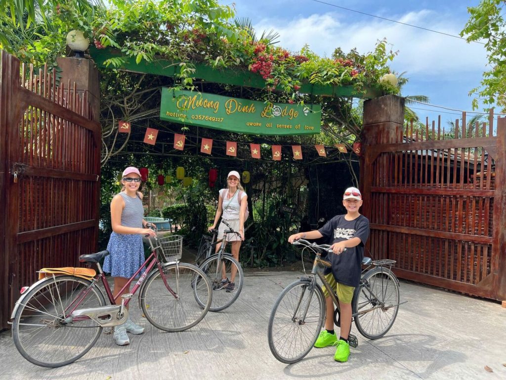 Two children and a woman with bicycles in front of the lodge's open wooden gates.