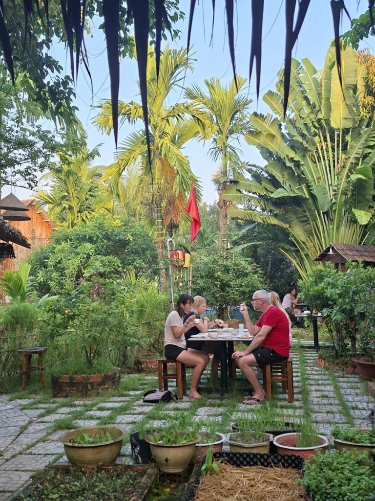 Guests eating at a table in a garden courtyard among potted plants and palms.