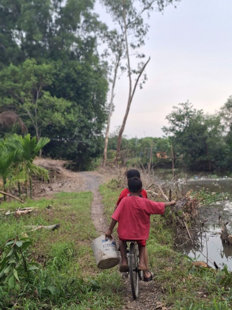 Two children in red T-shirts riding one bicycle along a grassy canal-side path, the rear one holding a bucket.