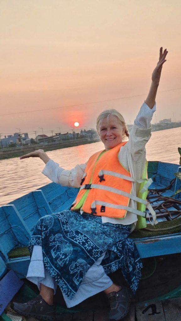 A woman in a life jacket raising her arms in a boat, the sun low over the river.