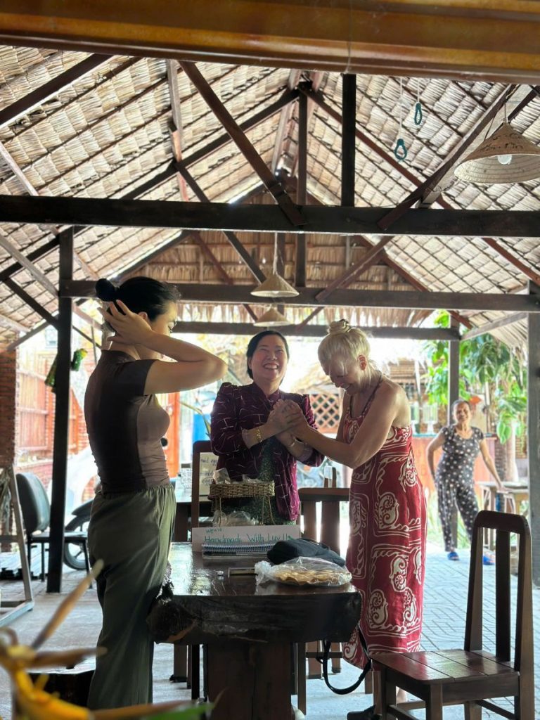 Three women chatting at a table under a large thatched pavilion.