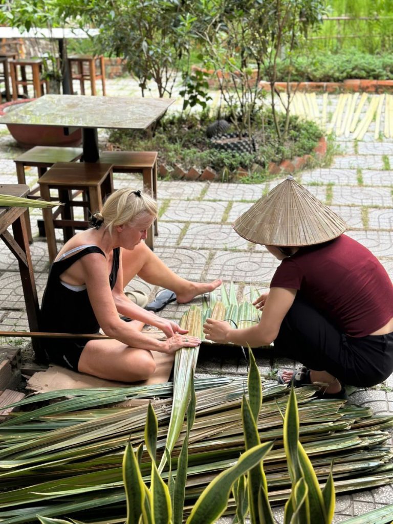 A guest and a woman in a conical hat weaving palm fronds on the ground.