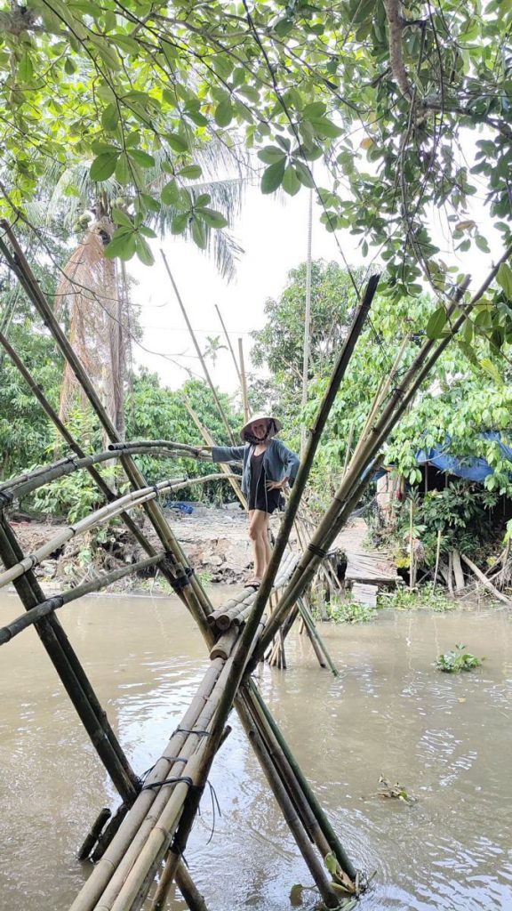 A person crossing a bamboo monkey bridge over a wide brown canal.