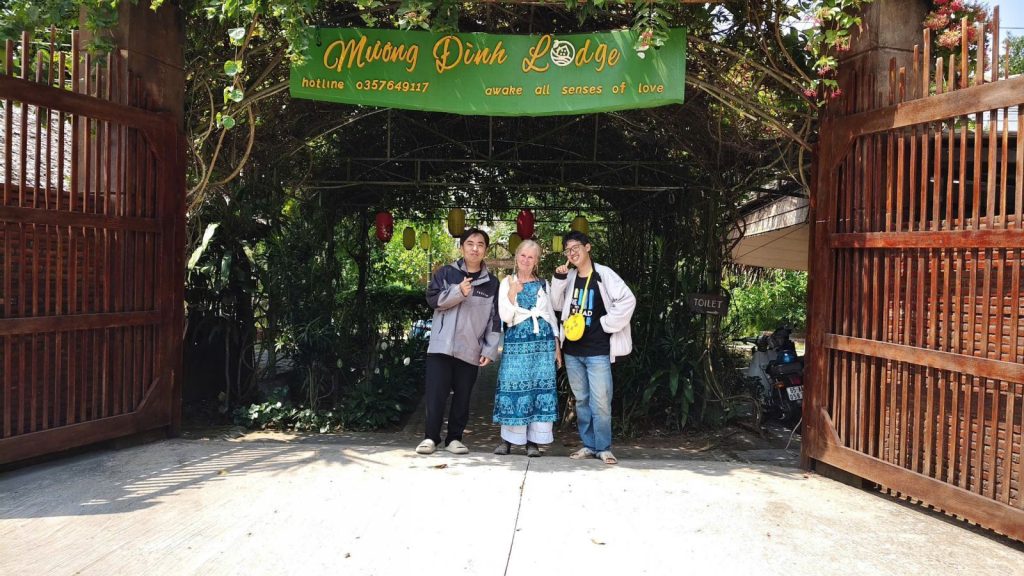 Three guests standing under the Mương Đình Lodge sign between open wooden gates.