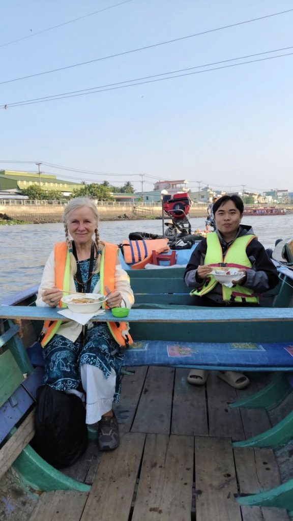 An older woman and a young man in life jackets seated in a boat with bowls of food, a town across the river.