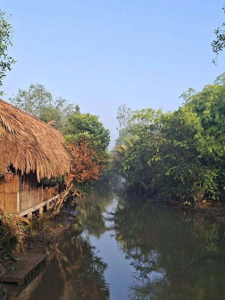 A still canal reflecting the sky between a thatched hut and green trees.