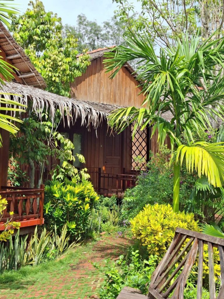 A thatched wooden cottage behind palms and flowering shrubs.