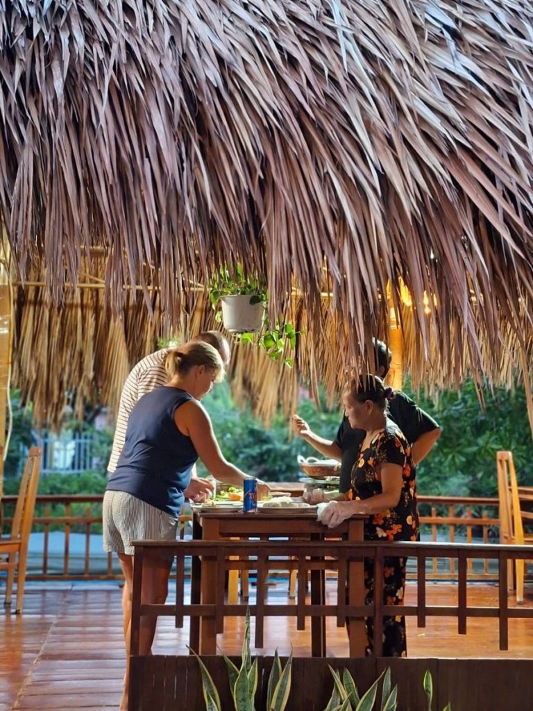 People preparing food at a table on a wooden deck under a thatched roof.