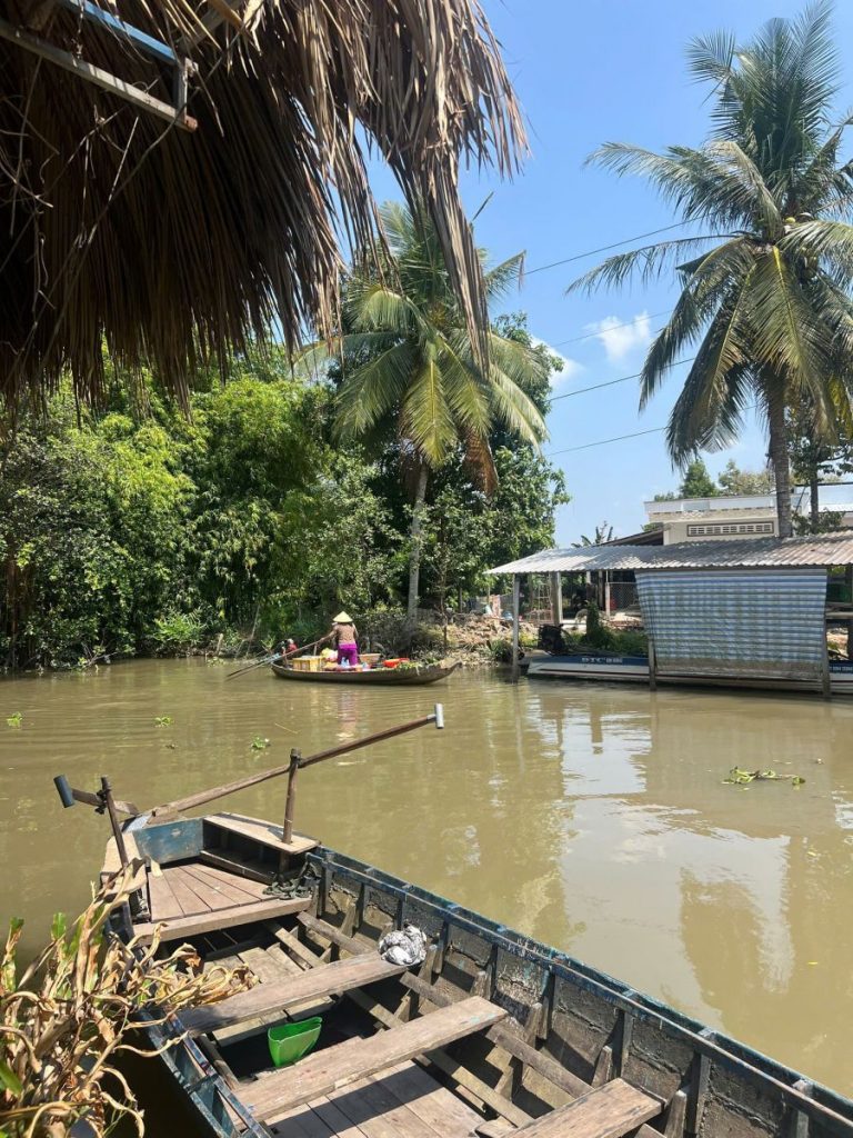 A wooden boat moored on a brown canal, with a small boat and a stilt house beyond.