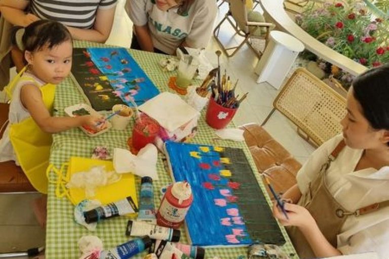Một em nhỏ vẽ tranh tại bàn workshop bên cạnh một phụ nữ, quanh bàn là cọ và tuýp màu.