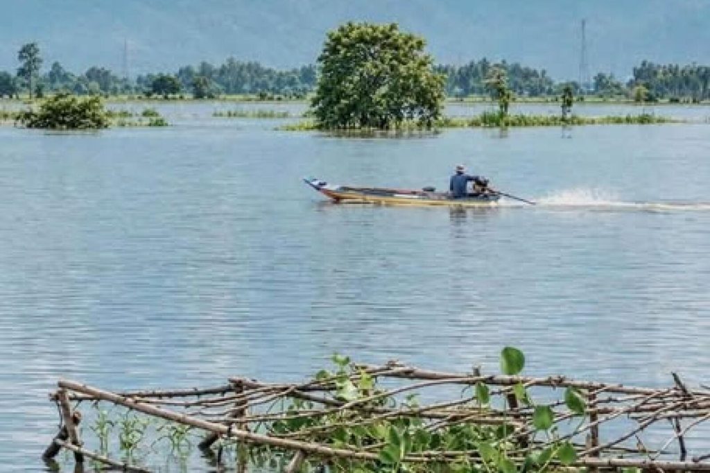 A man steers a long-tail boat across flooded fields, with trees rising from the water and hills in the haze.