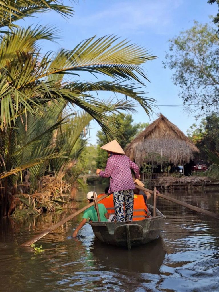 A rower in a conical hat takes passengers in life jackets along a canal.